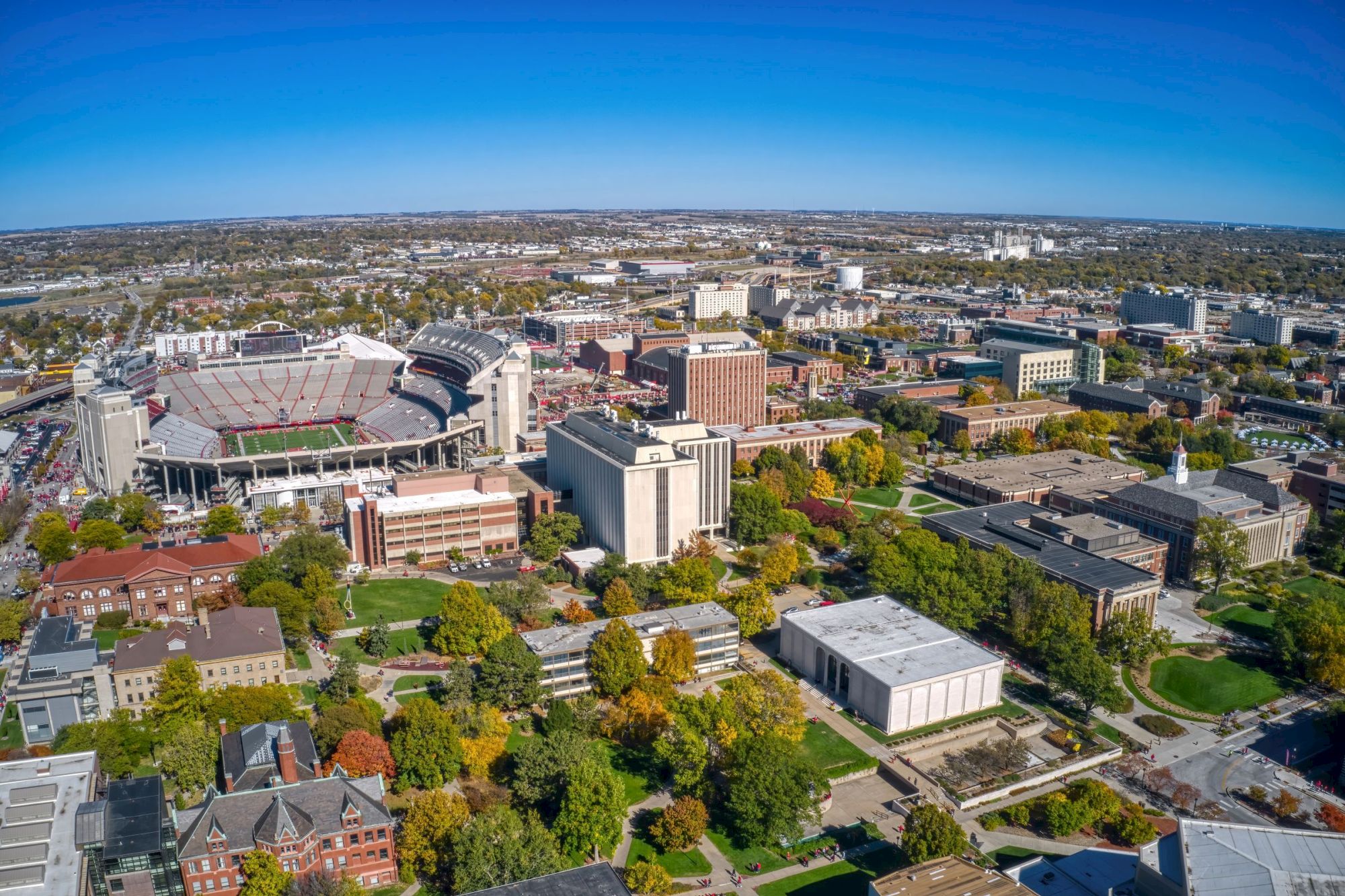An aerial view of a university campus with a large stadium, surrounding buildings, parking lots, and fall trees under a clear sky.