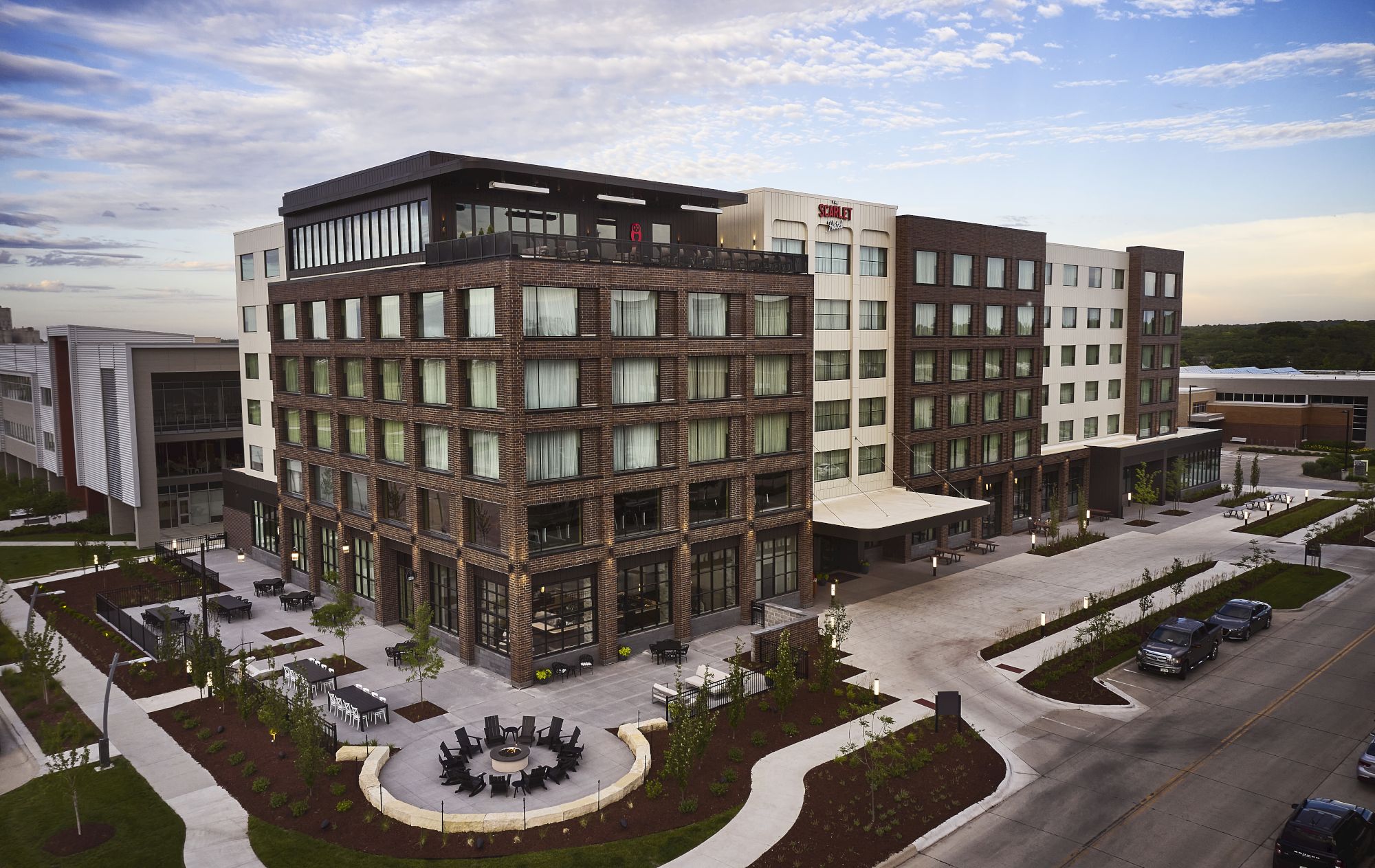 A modern hotel exterior with brick-and-glass rooms, a courtyard patio, lounge tables, and a bright blue sky above.