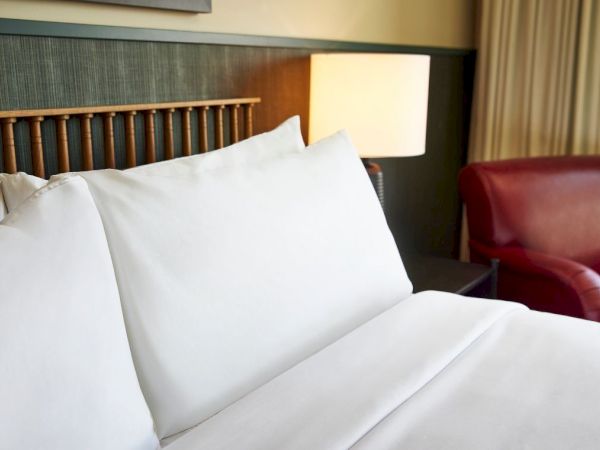 A neatly made bed with white pillows sits in a cozy hotel room, with a bedside lamp and a red leather chair in view.