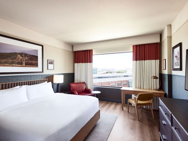 A modern hotel room with a large bed, red-and-white curtains, a sitting area, and a desk with a city view outside.