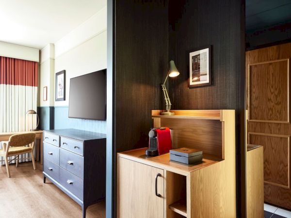A hotel-style room with a TV on a dresser, a chair by curtains, and a desk with a red espresso machine.