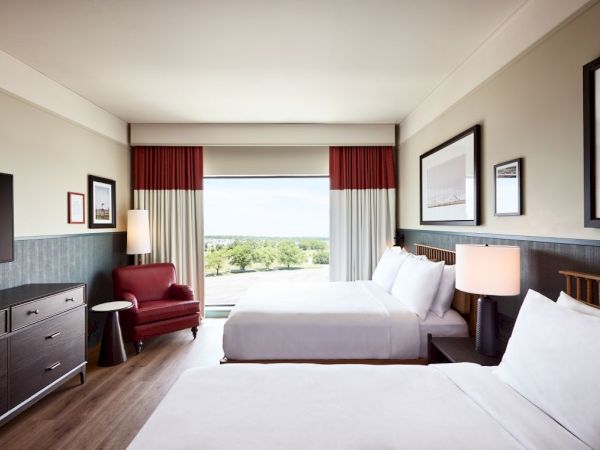 A modern hotel room with two white beds, red chair, lamps, framed art, and large windows with curtains.