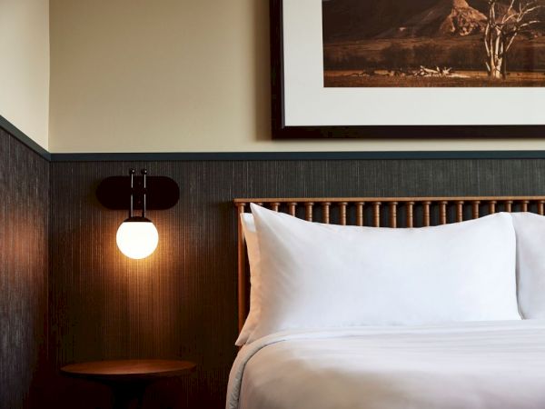 A hotel room corner with a neatly made bed, white pillows, a warm wall lamp, and framed landscape art.
