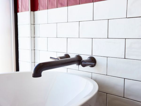 A dark metal bathroom sink faucet on a white tiled wall, with a hint of red curtain in the background.