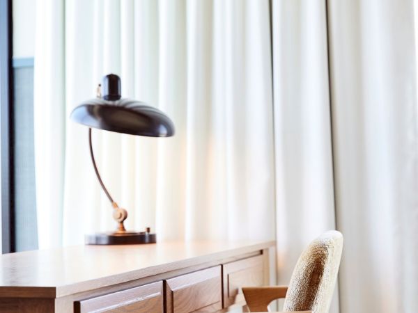 A small dark lamp on a wooden table beside a cushioned chair, with red-and-white curtains in the background.