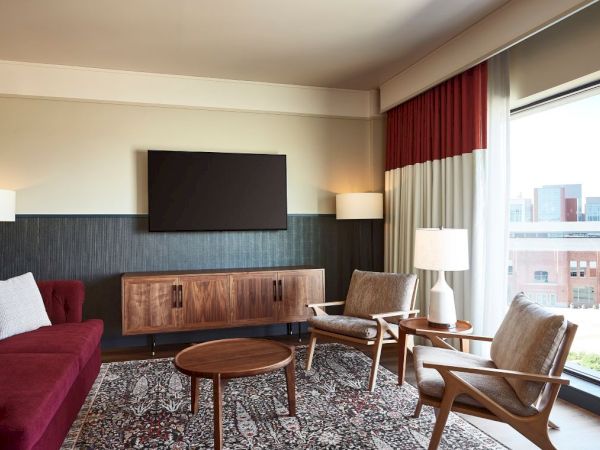 A modern hotel lounge with a TV on a wall, red sofa, coffee table, armchairs, floor rug, and tall window curtains.