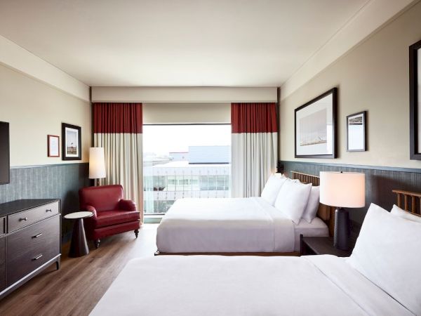 A bright hotel room with two beds, framed pictures, lamps, and red-and-cream curtains.