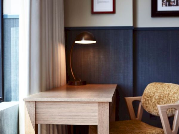A small office nook with a wooden desk, lit table lamp, and upholstered chair beside blue wall paneling.