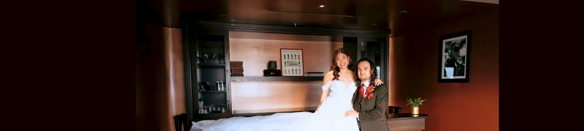 A couple in wedding attire poses in a modern room with warm lighting and shelving behind them.