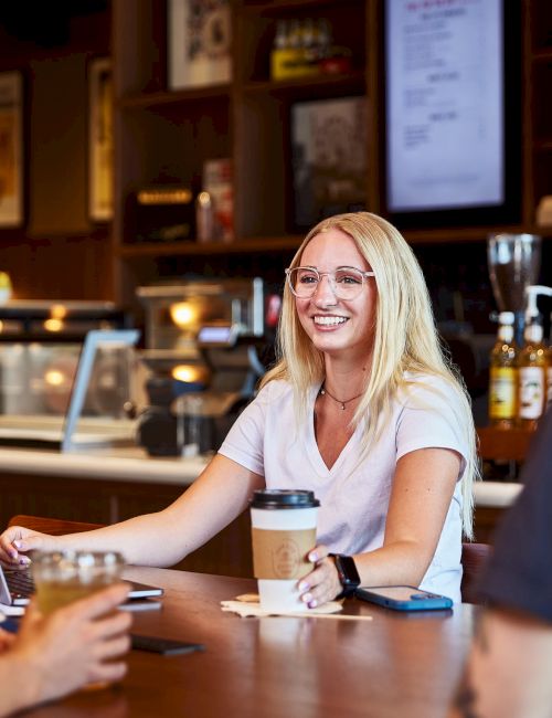 A blonde barista behind the counter in a cozy caf&eacute; chats with customers while holding a coffee, with laptops and drinks on the table.