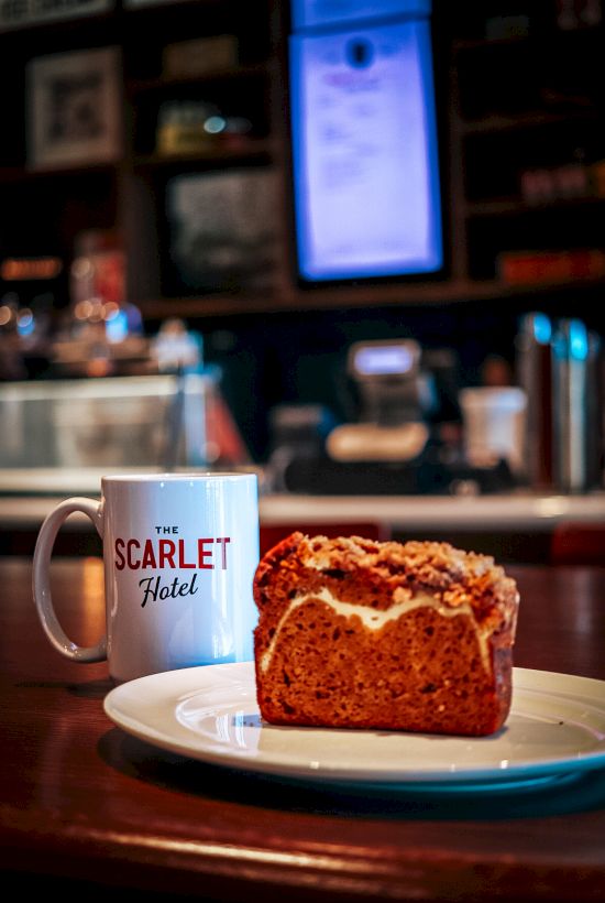 A caf&eacute; table shows a &ldquo;The Scarlet Hotel&rdquo; mug beside a slice of cake, with a blurred restaurant background behind it.