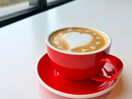 A red cup of latte with creamy foam and heart-like latte art on a white saucer by the window.