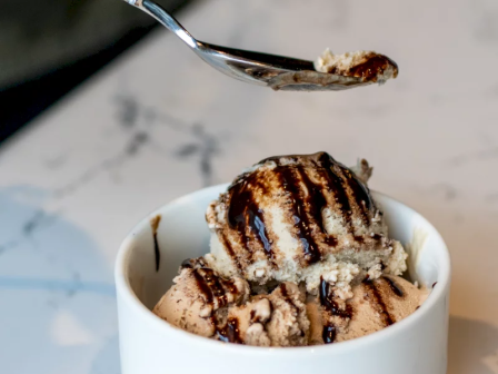 A spoon scoops chocolate-drizzled cookie dough or ice cream from a small white bowl.