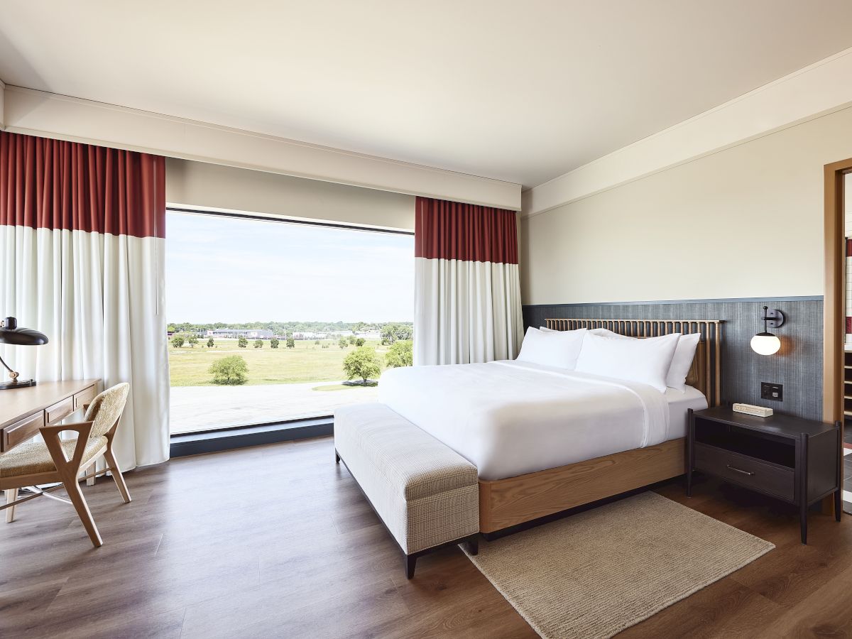 A modern hotel bedroom with a neatly made bed, red-and-white curtains, and a large window looking out onto a green field.