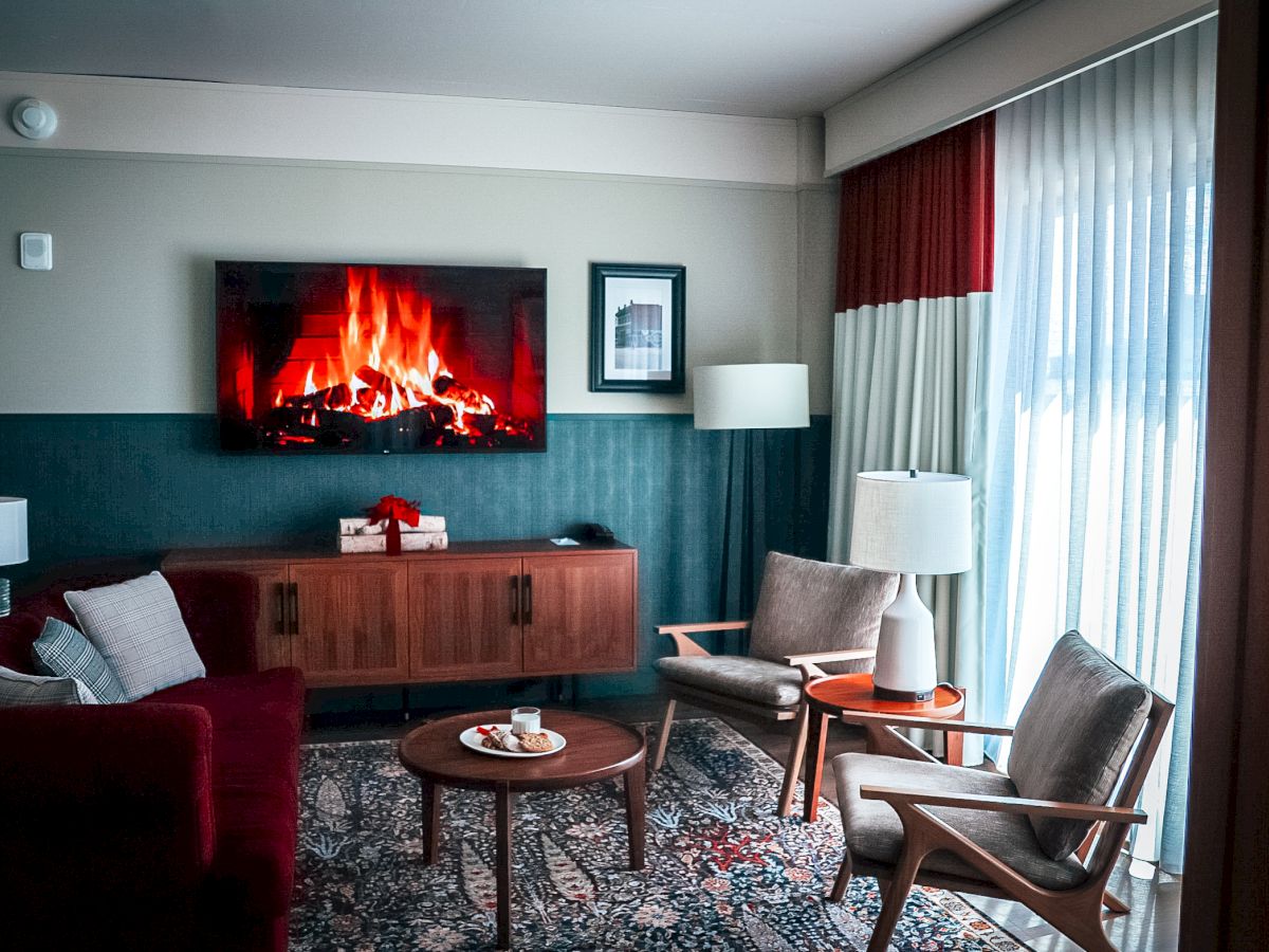 A cozy hotel living room with a glowing fireplace, sofa, armchairs, lamps, and a sideboard beside tall curtains.