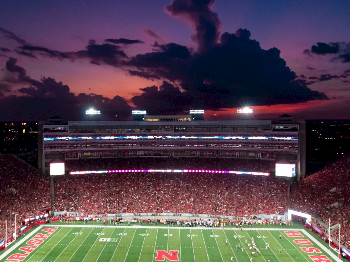 A packed football stadium at dusk with players on the field, bright lights, and a colorful smoke plume in the sky.