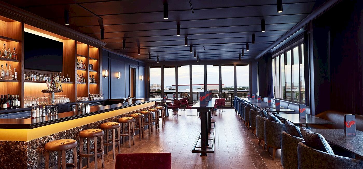 A sleek bar lounge with bottles, bar stools, plush seating, and floor-to-ceiling windows under pendant lights.