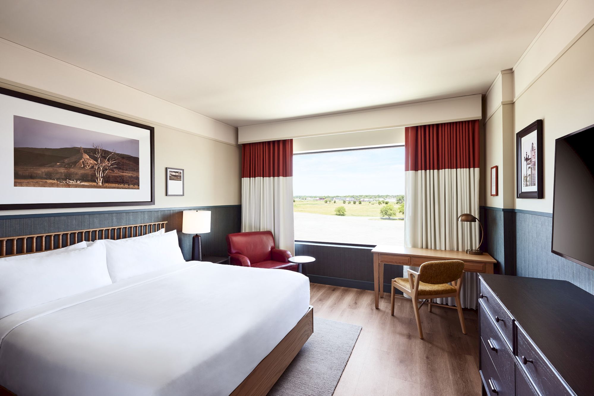 A bright hotel room with a large bed, red lounge chair, desk, and tall window curtains, overlooking open countryside.
