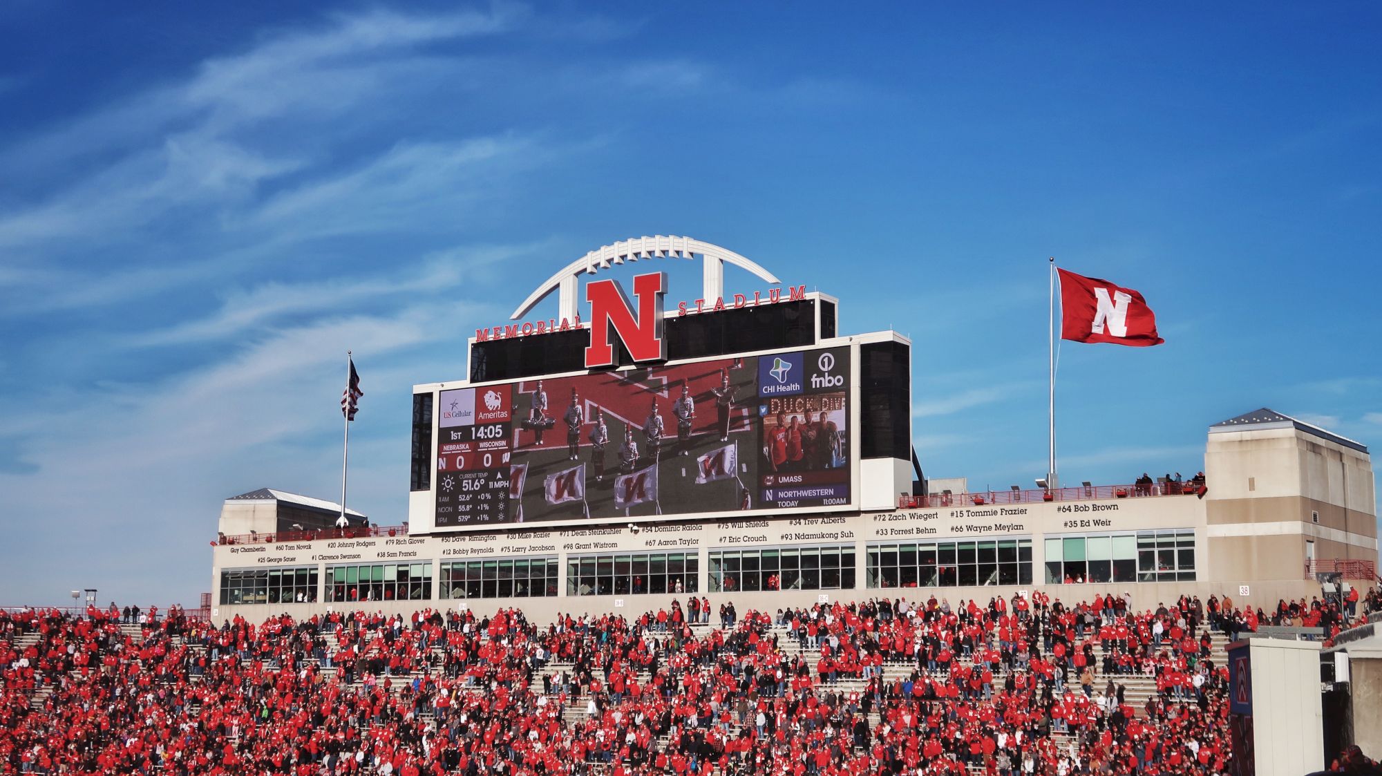 Fans in red fill Nebraska&rsquo;s Memorial Stadium as a giant N and flag fly above the scoreboard under a blue sky.