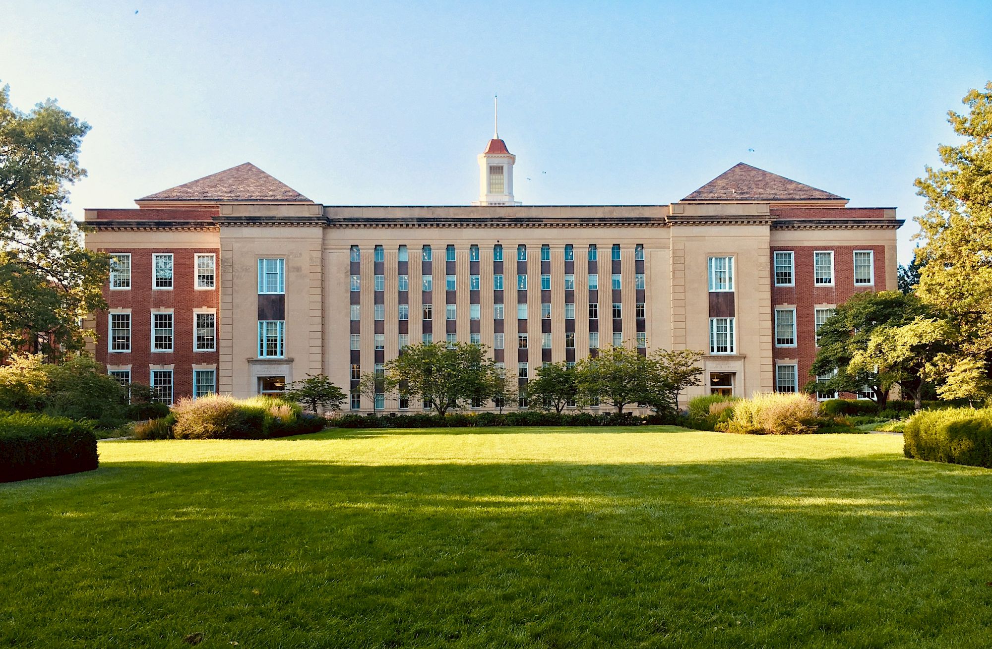 A grand brick campus building with tall columns and a white cupola rises above a manicured green lawn under blue sky.