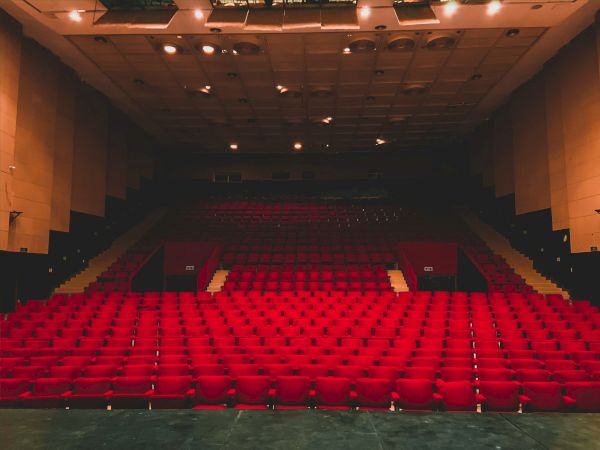 An empty auditorium with rows of red seats facing a dark stage under ceiling lights.
