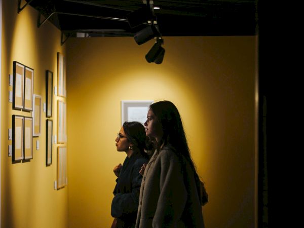 A hallway lit by spotlights, with two women standing in coats, looking toward a screen-like opening in the back.