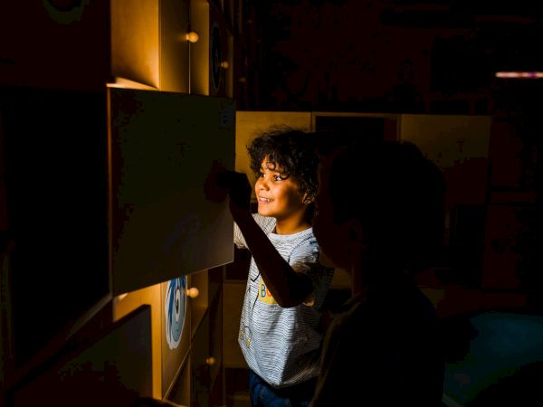 A child smiles while peeking out of a dark room, touching a wooden wall of lit toy drawers.