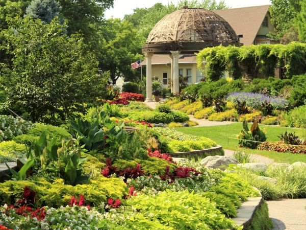 A lush flower garden leads to a white pavilion with a domed gazebo roof and an American flag waving.