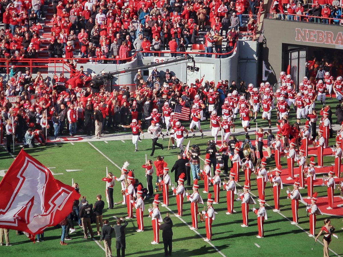 Nebraska football game: players and a huge crowd in red fill the field while the marching band lines up near the stadium tunnel.