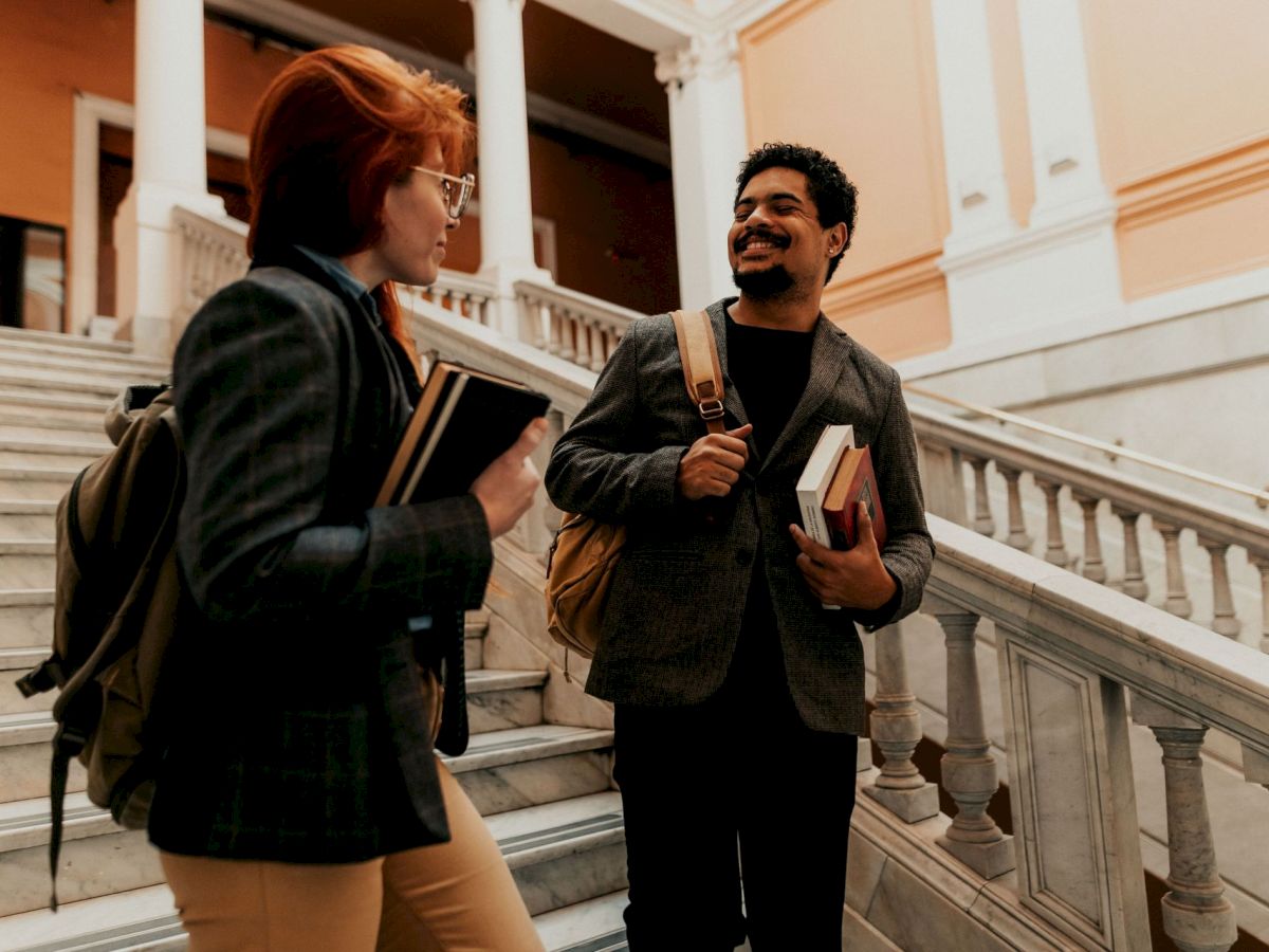 Two students chat on marble steps in a grand hall, one holding textbooks and smiling at the other on campus.