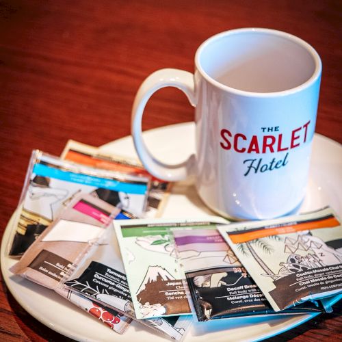A &ldquo;The Scarllet Hotel&rdquo; mug sits on a wooden table beside assorted tea and snack packets on a tray.