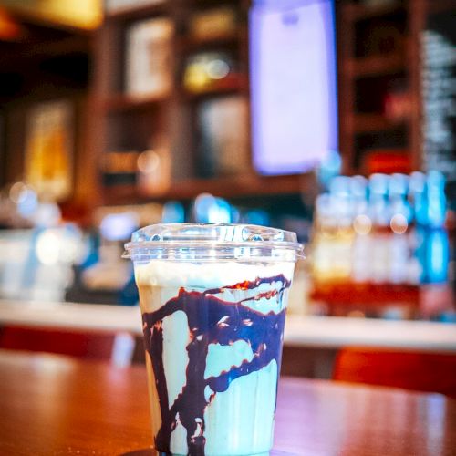 A plastic cup of dark-and-light ice cream or dessert sits on a table in a dim caf&eacute;/bar, with blurry drinks behind it.