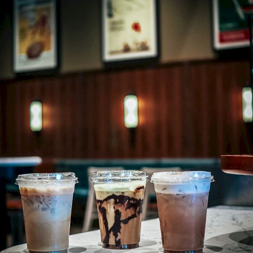Three iced coffee drinks in clear cups sit on a marble counter in a cafe, with blurred menu boards behind them.