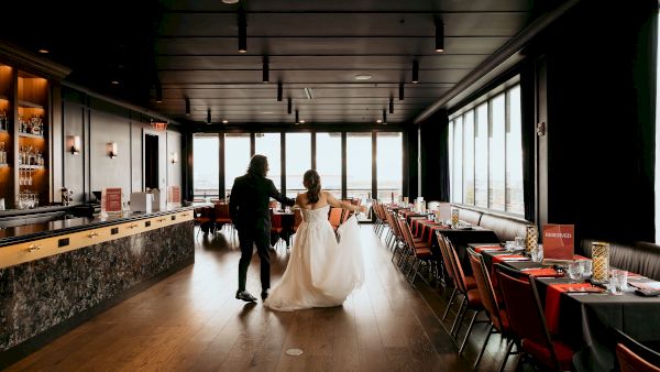A wedding couple in a modern restaurant, walking hand-in-hand beside a bar and rows of reserved tables.