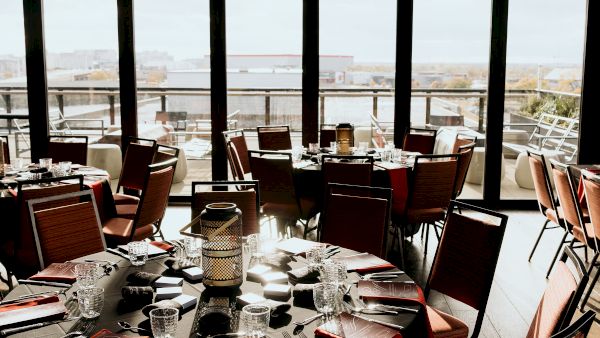 A sunlit restaurant dining area with set tables, glassware and chairs, overlooking a city view through large windows.